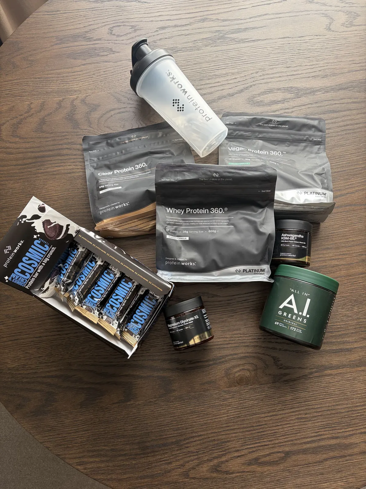 Protein Works Clear Protein 360, Whey Protein 360 PLATINUM and Vegan Protein 360 PLATINUM bags on a table with a Protein Works shaker